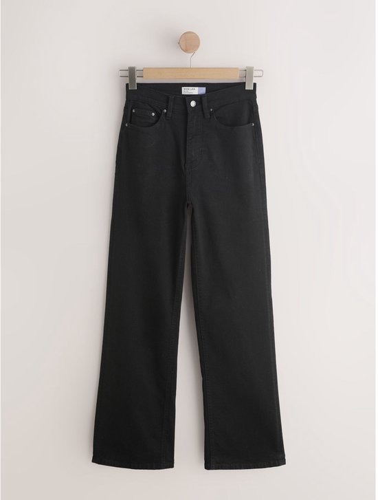 Zwarte jeans met wijde pijpen hangend aan een houten kledinghanger tegen een lichte muur.