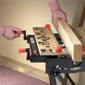 Een hand met een oranje handvat draait aan de werkbank, terwijl een andere hand een houten plank vasthoudt.