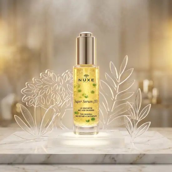 Nuxe Super Serum [10] anti-age serum in glazen flesje met gouden dop, omringd door decoratieve bladeren.