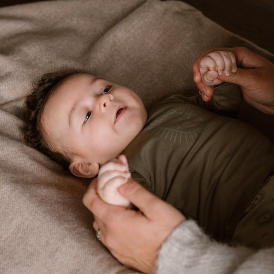 Close-up van een baby in een kaki romper met een leeuwenprint, die de hand van een volwassene vasthoudt.
