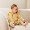 Baby in geel geruit jurkje met kanten kraagje en bloemenprintje op de borst, zittend op een bank.