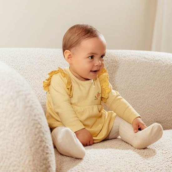 Baby in geel geruit jurkje met kanten kraagje en bloemenprintje op de borst, zittend op een bank.