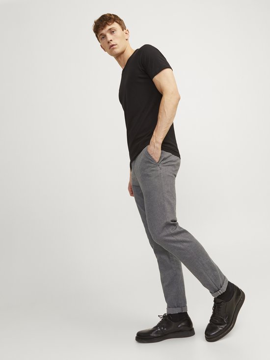 Model draagt een zwarte t-shirt en een grijze broek met fijne structuur, met de hand in de zak.