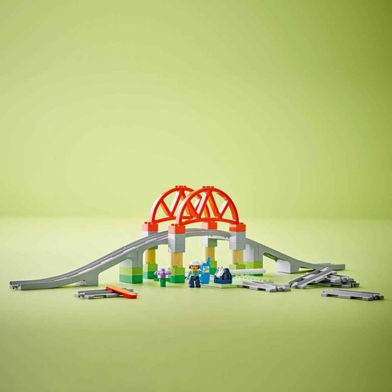 Brug van oranje en grijze blokken, met groene en beige steunen. Spoorrails en kleine figuren.