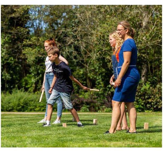 Vier mensen spelen Kubb in een zonnig park; een jongen werpt een stok.