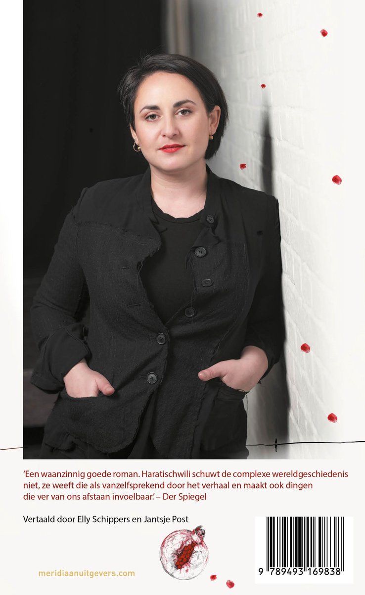 Vrouw met kort zwart haar en rode lippen, staand voor een witte bakstenen muur, met een zwart vest aan.