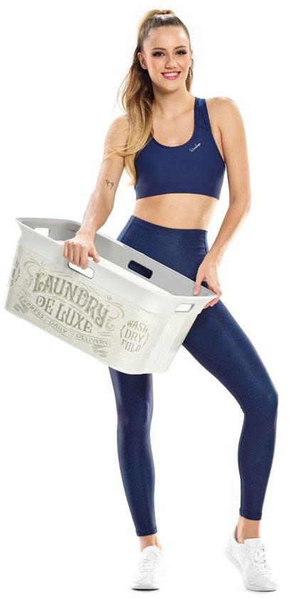 Model in navy sportbeha en legging, houdt een witte wasmand vast, met witte sneakers.