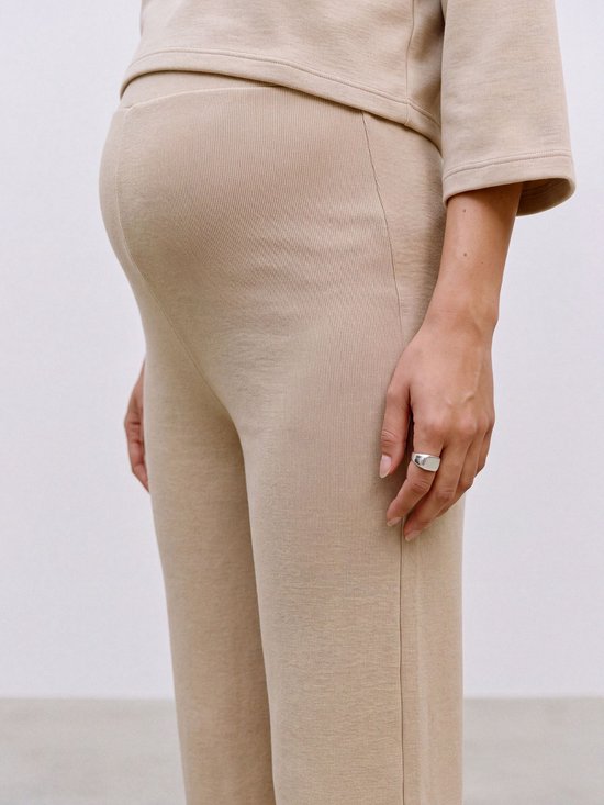 Close-up van een zwangere buik, gedragen met een beige geribbelde broek met wijde pijpen en een bijpassende top. Een hand met een zilveren ring is zichtbaar.