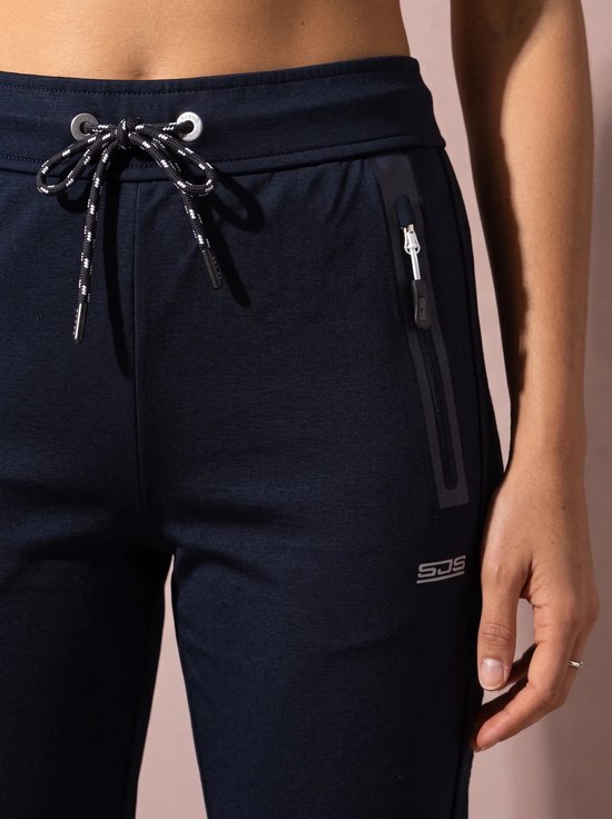 Detailopname van de marineblauwe sportbroek met trekkoord, ritszakken en SJS-logo.
