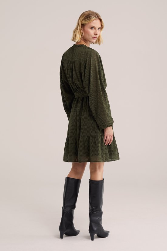 Dames jurk in donkergroen met een strikceintuur, lange mouwen en een gelaagde rok. Gekleed met zwarte laarzen.