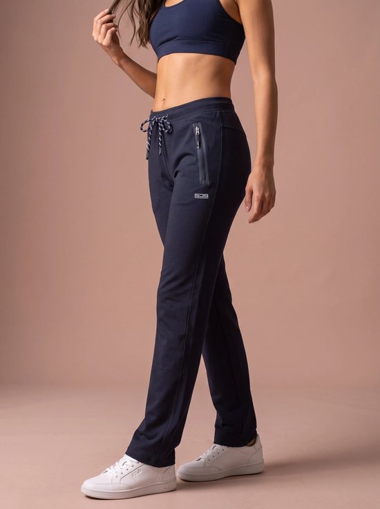 Zijaanzicht van een model in een marineblauwe Sjeng trainingsbroek met ritszakken en witte sneakers.