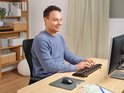 Man in een blauwe trui typt op een Trust Ody II draadloos toetsenbord aan een houten bureau.