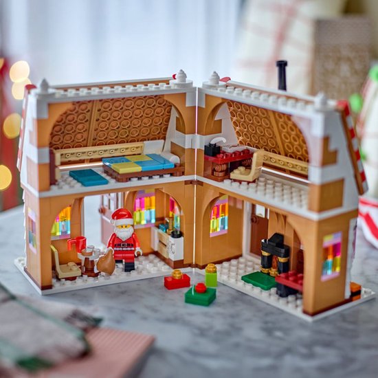 Een open LEGO peperkoekhuisje met een kerstman met een zak. Binnen zijn een bed, keuken, piano en een bureau te zien.
