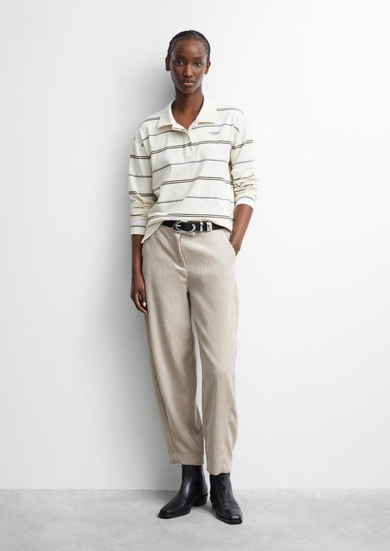 Model in gestreept crème poloshirt met 'comma' logo en beige corduroy broek, gecombineerd met een zwarte riem en laarzen.