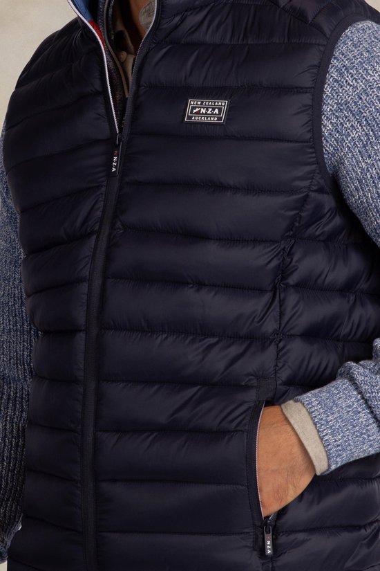 Close-up van donkerblauwe gewatteerde bodywarmer met Patch 'New Zealand Auckland', gedragen over een blauw gebreid vest.