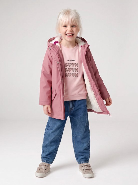 Meisje met blond haar, lachend, draagt een roze regenjas met teddy voering, roze trui met hartjesprint en spijkerbroek.