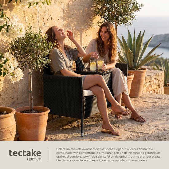 Twee lachende vrouwen genieten van drankjes op een zwarte polyrattan tuinbank met tafel op een zonnig terras.