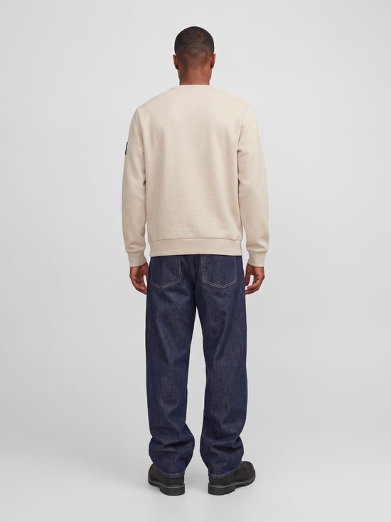 Zijaanzicht van de achterkant van een man in een crème kleurige sweater en donkerblauwe jeans. De sweater heeft lange mouwen en een label op de mouw.