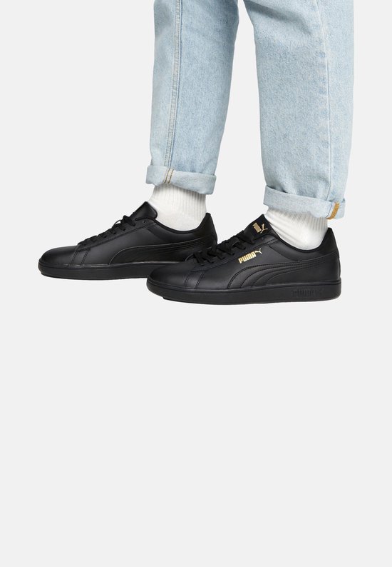 Voeten in lichtblauwe spijkerbroek en witte sokken, met zwarte Puma Smash sneakers.