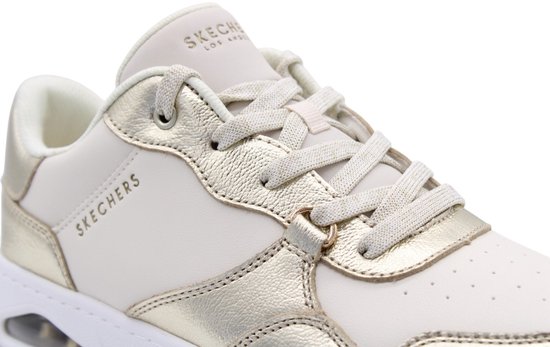 Close-up van een champagnekleurige Skechers sneaker met veters, metalen accenten en het merklogo.