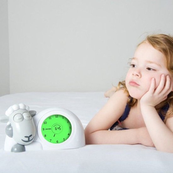 Een meisje ligt op bed naast een witte schaapwekker met groene wijzerplaat. De wekker toont 6:45.