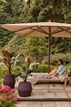 Vintage pruimkleurige Elho bloempot op een terras met een vrouw die onder een parasol op een ligbed ontspant.