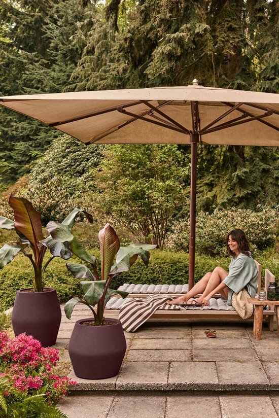 Vintage pruimkleurige Elho bloempot op een terras met een vrouw die onder een parasol op een ligbed ontspant.
