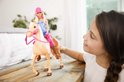 Kind kijkt naar een Barbie-pop op een paard met een roze helm in een lichte kamer.