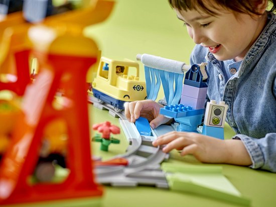 Kind speelt met een gele trein en grijze rails, met een blauwe wasstraat op de achtergrond.