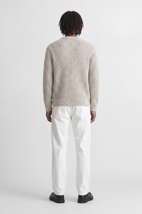 Model in beige grofgebreide trui en witte broek, volledig zicht van achteren.