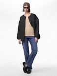 Model draagt een beige hemdje, een zwarte doorgestikte jas, blauwe jeans, zonnebril en pet.