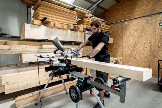 Man met baard, zwarte polo, tatoeages op armen, zaagt houten balk met Metabo afkortzaag KGS254M.