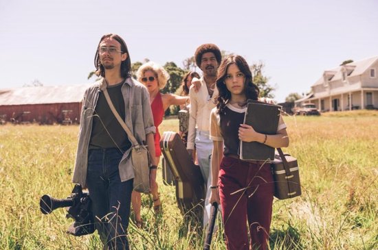 Groep van vijf mensen loopt door een zonnig veld, sommigen met koffers en een camera. Inclusief Jenna Ortega.