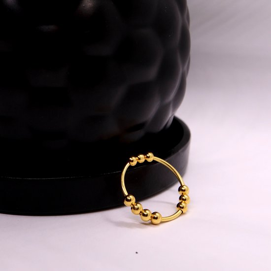 Gouden ring met kleine balletjes, geplaatst op een zwarte, abstracte vorm.