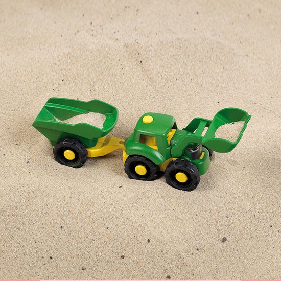 Een groene en gele John Deere speelgoedtractor met aanhanger, liggend in zand.