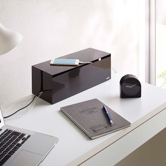 Boîte à câbles gris foncé sur un bureau, avec un smartphone posé dessus, à côté d'un ordinateur portable et d'un carnet.