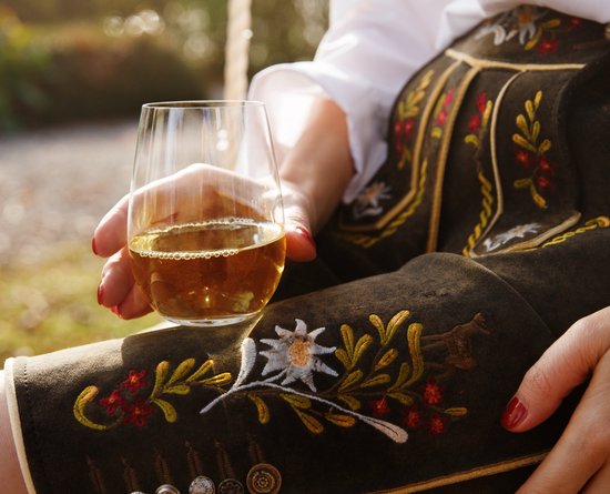 Hand houdt een Riedel O Wine Tumbler met witte wijn vast. Vrouw draagt traditionele Beierse klederdracht.