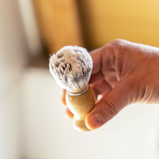 Hand houdt een scheerkwast vast met bamboe handvat en bruin-witte haren.
