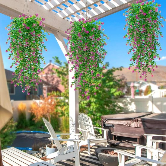 Drie hangende planten met roze bloemen en groene bladeren, onder een pergola in een tuin met tuinmeubilair.