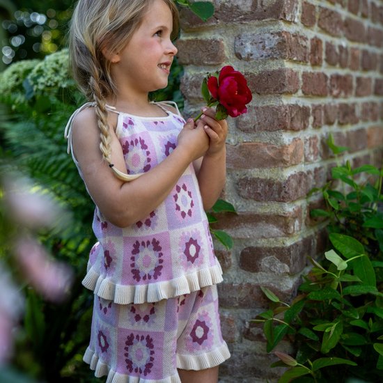 Lachend meisje met blonde vlechtjes in lila top met bloemenpatroon en ruches, houdt een rode bloem vast.