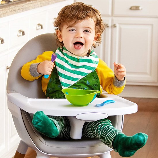 Een vrolijke baby zit in een kinderstoel met een slabbetje en eet met een blauwe vork.
