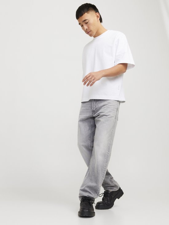 Man met witte T-shirt en grijze jeans, kijkend naar de vloer.