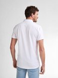 De achterkant van een witte polo met een klein logo op de linkerborst, gedragen door een man met een lichte spijkerbroek.