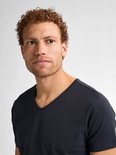 Close-up van een man met krullend bruin haar en sproeten, die een donkerblauw V-hals T-shirt draagt.