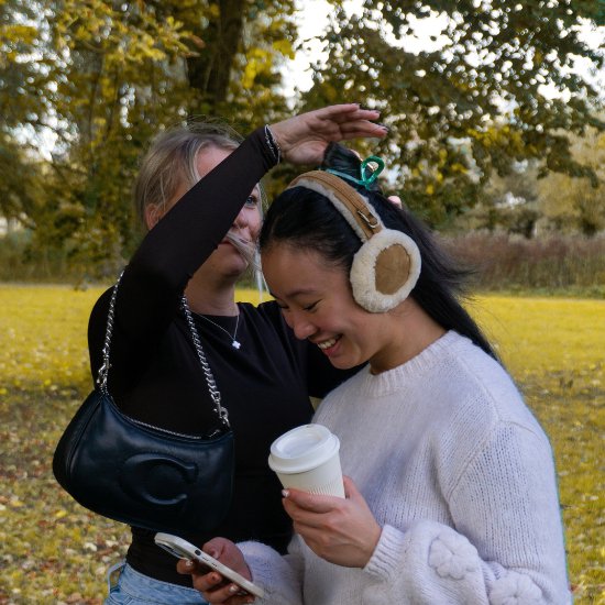 Vrouw met oorbeschermers en een groene haarklem in haar haar, lachend terwijl een andere vrouw haar haar aanraakt.