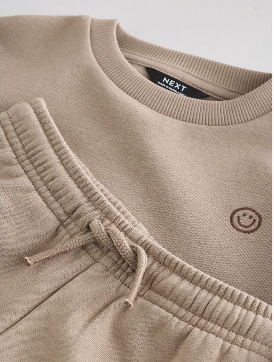 Detailopname van het NEXT-label in de naturelkleurige sweater met lachend gezichtje en joggingbroek.