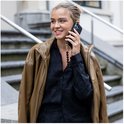Vrouw met bruine jas en zwart shirt, aan de telefoon, met een zwart telefoonhoesje met een hartjesketting.