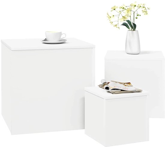 Drie witte kubusvormige bijzettafels, één met een kopje koffie, één met tijdschriften, en één met een vaas bloemen.