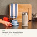 Beige melkopschuimer met een blauw gloeiend aan-uit icoon. Een hand pakt een lichtblauw schoteltje met kopje op. Een rode kop staat ernaast.