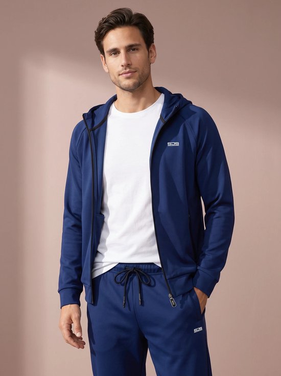 Man met donker haar draagt een donkerblauw vest met rits en capuchon met wit T-shirt en bijpassende broek. Logo op vest en broek.